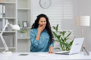 sobrecargado de trabajo mujer de negocios con Rizado pelo sensación exhausto, bostezando a su moderno oficina espacio con ordenador portátil y oficina suministros. foto