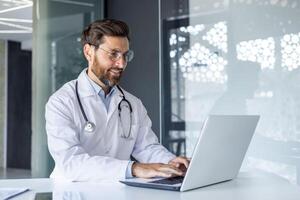 un sonriente joven masculino médico es trabajando en el oficina de el clínica utilizando un ordenador portátil. sentado en un blanco Saco a el escritorio, mecanografía, enviar mensajes de texto de cerca foto. foto