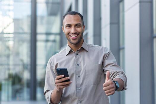 Young African American businessman outside office building smiling and looking at camera, thumbs up, programmer using smartphone for app testing photo