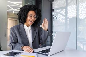 africano americano joven mujer de negocios, diseñador trabajando en oficina a computadora portátil, saludo y hablando en línea en llamar. foto