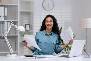 un enfocado Hispano mujer análisis documentos en su bien organizado hogar oficina, exhibiendo negocio eficiencia. foto