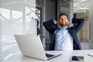 contento y sonriente joven empresario hombre sentado en el oficina a el escritorio y descansando y satisfecho ,con el trabajo con su manos detrás su cabeza. foto