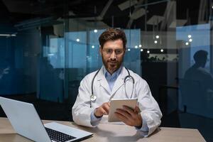 maduro médico en un médico vestido es trabajando dentro clínica oficina, médico es participación un tableta computadora en su manos para en línea consulta de pacientes, trabajando con un ordenador portátil. foto
