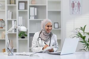un alegre musulmán hembra médico vistiendo un hijab se involucra en un en línea consulta, tomando notas y sonriente en un bien organizado médico oficina. foto