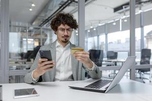 Focused young businessman with curly hair multitasking in a modern office, working on a laptop while checking his phone. photo