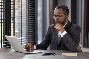 un profesional empresario en un traje concentrando en su trabajo en un ordenador portátil a un moderno oficina escritorio, posiblemente analizando datos o planificación estrategias. foto