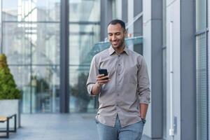 Successful young african american man walking outside office building, engineer software developer programmer smiling and happy using test app on phone, happy satisfied with result photo