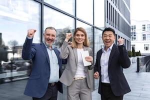 Successful diverse business team, three loving men and woman looking at camera and happy celebrating victory, team dreaming outside office building, business group in business suits photo