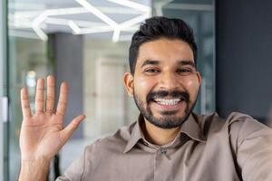 Cheerful businessman engaging in a call while waving and smiling at the camera, set in a modern office environment. photo