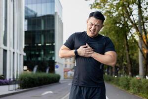 joven asiático hombre haciendo Deportes a el estadio. él es en pie doblado encima, participación su mano a su pecho, sufrimiento desde dolor, corazón ataque, cansancio excesivo. foto