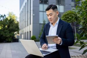 grave joven hombre, asiático empresario trabajando fuera de un oficina edificio utilizando un ordenador portátil y haciendo notas en un cuaderno. foto