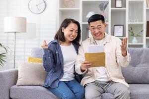 alegre y contento asiático familia juntos, hombre y mujer a hogar en sofá sentado en vivo habitación, recibido postal sobre con bueno noticias, mensaje y bueno logro noticias. foto