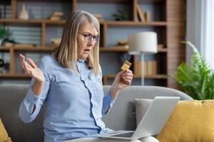 Mature woman sitting on the couch looking puzzled with a credit card and a laptop, possibly dealing with online shopping or banking issues. photo