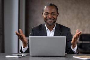 satisfecho empresario mirando felizmente a portátil computadora pantalla y extensión manos mientras sonriente ampliamente. oscuro peludo hombre sensación satisfecho mientras viendo resultado de diario difícil trabajo a hogar oficina. foto