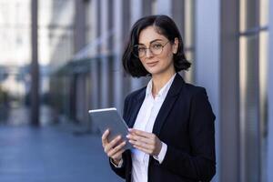 Portrait of a self-confident business woman, executive director, company founder standing in a suit and glasses near the office center, holding a tablet in her hands and smiling at the camera. photo