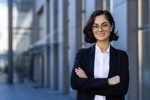 Young beautiful businesswoman in business suit smiling and looking at camera, portrait of successful woman outside office building, female worker in glasses with crossed arms. photo