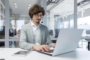 Focused young adult male entrepreneur with curly hair using laptop and headset in a modern office setting, embodying professionalism and dedication. photo