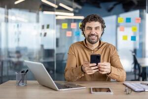 Curly caucasian man sitting in cabinet with paper stickers on walls and looking at camera with sincere smile. Adult male in shirt using set of digital gadgets for practising multitasking at work. photo