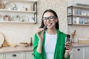 joven hermosa morena mujer en lentes y verde camisa a hogar en cocina, comiendo chocolate foto