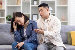 familia conflicto, psicológico violencia, abuso. joven asiático hombre y mujer son discutiendo mientras sentado en el sofá a hogar. el mujer escucha y sostiene su cabeza, el hombre gritos y amenaza foto