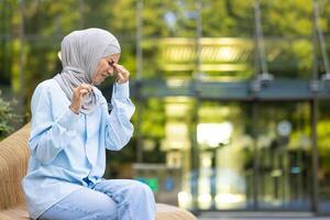 adulto musulmán mujer en azul hijab experimentando grave dolor de cabeza o migraña, participación cabeza en dolor mientras sentado fuera de en un banco. foto