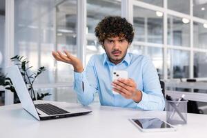 Worried young hispanic man sits in the office at the desk and looks disappointedly at the phone, spreads his hands, looks upset at the camera. photo