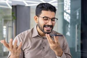 sonriente joven adulto masculino se involucra en un alegre conversacion utilizando un manos libres móvil teléfono en un moderno oficina ajuste. foto