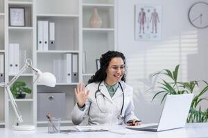 Friendly Hispanic female doctor in white coat waving during a call consultation in a bright office setting. photo