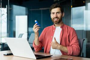 Joyful man with inhaler in hands smiling and looking at camera, showing thumbs up recommending asthma medicine to ease breathing, working inside office at workplace. photo
