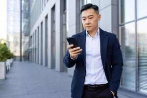 grave joven asiático hombre en pie fuera de un oficina edificio en un traje, participación su mano en su bolsillo y utilizando un móvil teléfono. foto