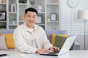 retrato de exitoso asiático programador, hombre trabajando remotamente desde hogar oficina, empresario sonriente y mirando a cámara utilizando ordenador portátil a trabajo sentado en vivo habitación a hogar. foto