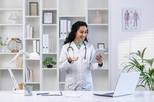 Doctor inside the medical office stands with a tablet computer in hands, a clinic worker in white medical robe will advises the patient remotely using the for call, joyfully and friendly. photo