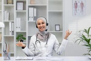 sonriente hembra médico vistiendo un hijab y auriculares comunica con un paciente vía llamar, sentado en un bien organizado médico oficina. foto