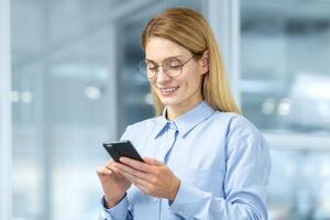 Portrait of attractive blonde woman in round eyeglasses looking down at cell phone in hands and smiling. Caucasian female making break from business and checking social media through digital gadget. photo