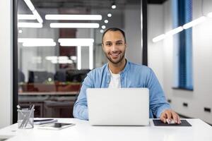 retrato de un confidente masculino profesional trabajando en un ordenador portátil en un bien iluminado contemporáneo oficina espacio, mirando a cámara. foto