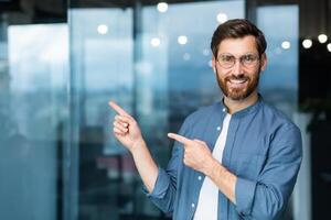 Smiling businessman inside office pointing fingers to direction, happy boss in shirt and glasses. photo