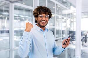 Businessman closeup in modern office, portrait successful man celebrating victory and good achievement results, hispanic smiling and looking at camera holding smartphone, receiving good news online. photo