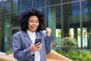 Close-up photo of young African American woman in suit standing near office center, holding and looking at mobile phone, happy showing victory gesture with hand.