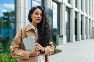 A confident, curly-haired professional woman stands outside a contemporary office building, holding a tablet and looking pensive. She appears focused and ready for business in a stylish outfit. photo