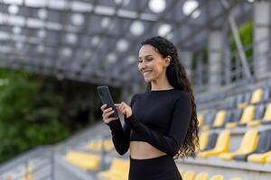 un ajuste mujer en ropa de deporte sostiene un teléfono inteligente mientras en pie en un estadio, exhibiendo un moderno y activo estilo de vida. foto