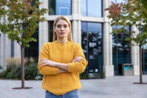 un pensativo mujer con doblada brazos vistiendo un vibrante amarillo suéter soportes con confianza en un urbano al aire libre configuración, transporte profesionalismo y determinación. foto