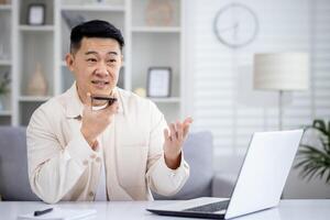 Young asian man sitting at home at table and working remotely on laptop, talking on speaker phone, using voice search. photo