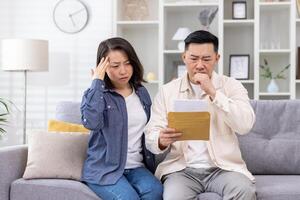 Upset Asian family, man and woman sitting together at home in living room on sofa, couple received letter mail envelope notification with bad news, unhappy with received message. photo