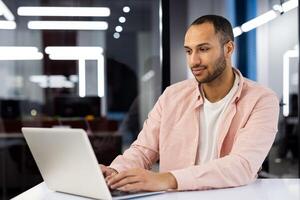 A young hispanic man is sitting in a modern office at a desk in a pink shirt, working and studying on a laptop. photo