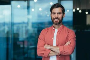 Portrait of a happy and smiling programmer in the middle of a modern office, a man in glasses and a red shirt is looking at the camera with his arms crossed. photo