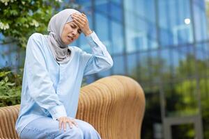 Stressed islamic woman having seat on bench while struggling with severe migraine outside. Exhausted female in cotton shirt grabbing head while feeling tensed and tired of high blood pressure. photo