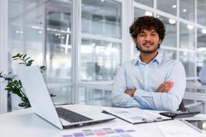 Portrait of mature businessman with beard, man in shirt smiling and looking at camera sitting at desk with crossed arms inside modern office building, using tablet and laptop at work. photo