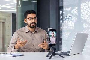 Successful business trainer teacher recording online training course, man inside office at workplace using phone and tripod, sitting at desk with laptop. photo