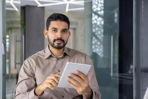 Confident young adult male with a beard, using a digital tablet in a well-lit, modern office environment. Perfect for depicting professionalism and technology in the workplace. photo