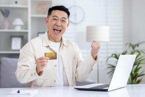 Close-up portrait of a young Asian man sitting at home at a table with a laptop, holding a credit card, and looking happy at the camera, showing a victory gesture with his hand. photo